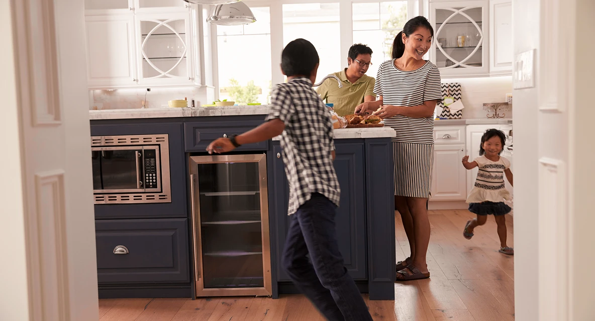 Family in a modern kitchen, with mother preparing food, father nearby, and two children playing and running.